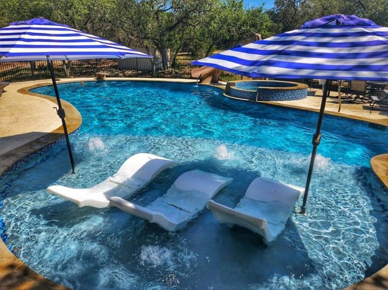 3 loungers partially submerged in a pool with 2 nearby umbrellas, hot tub seen in the background.
